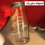 بانکه شیشه ای استوانه سایز یک در دست با بک گراند قرمز و درب فلزی